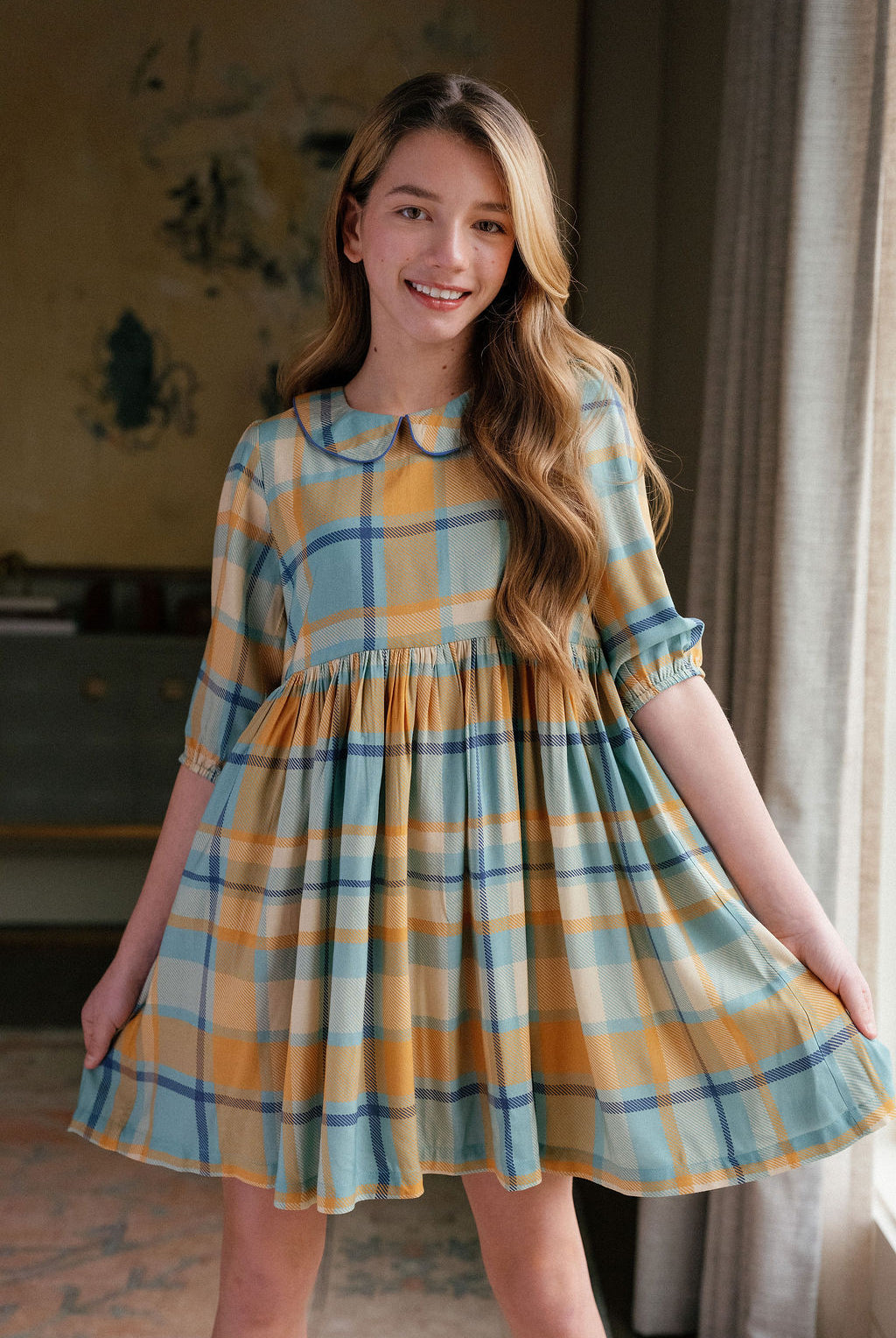 Girl wearing the Briton Court Annabel Plaid Dress featuring a classic blue and gold plaid print, Peter Pan collar with contrast piping, empire waist, and gathered skirt.