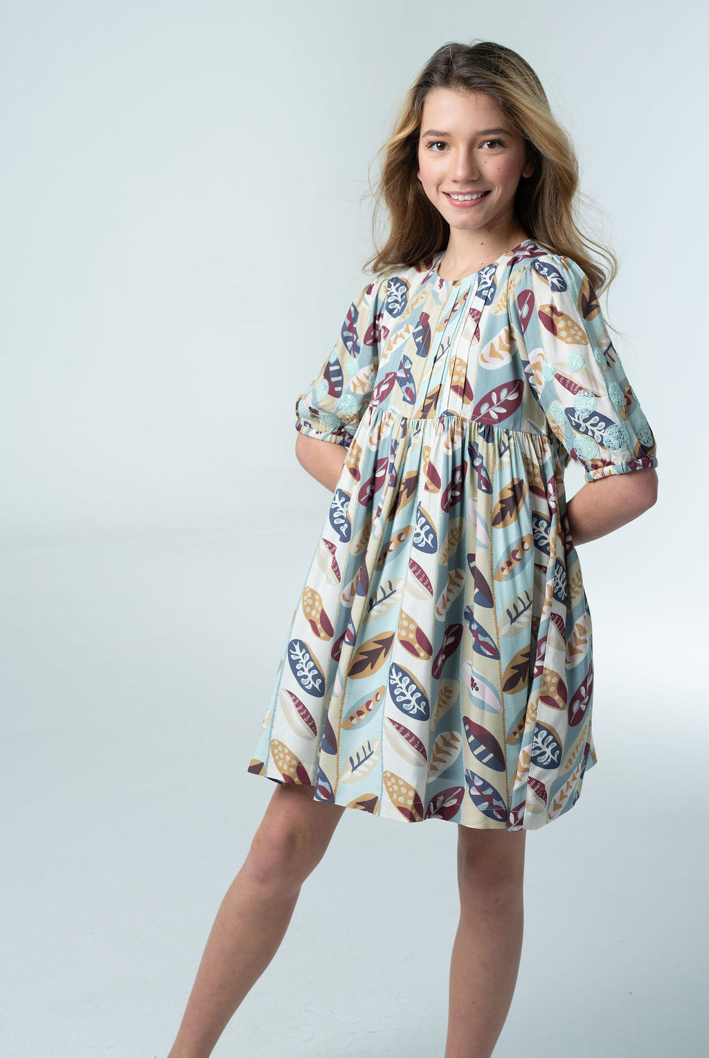 Girl wearing the Briton Court Eva Dress in the Falling Leaves print, featuring an allover leaf pattern in blue, burgundy, mustard, and sage with an A-line silhouette and puff sleeves.