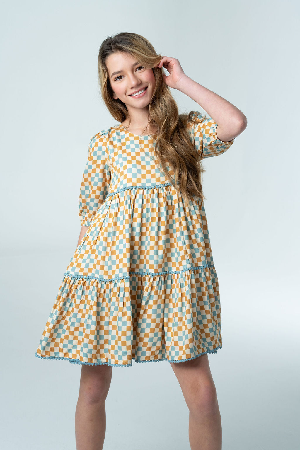 Girl wearing the Briton Court Quinn Tiered Checker Dress featuring a vintage-inspired gold and blue checker print with tiered skirt, blue contrast trim, puff sleeves, and hidden pockets.