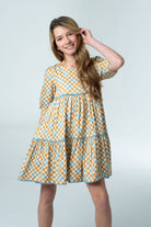 Girl wearing the Briton Court Quinn Tiered Checker Dress featuring a vintage-inspired gold and blue checker print with tiered skirt, blue contrast trim, puff sleeves, and hidden pockets.