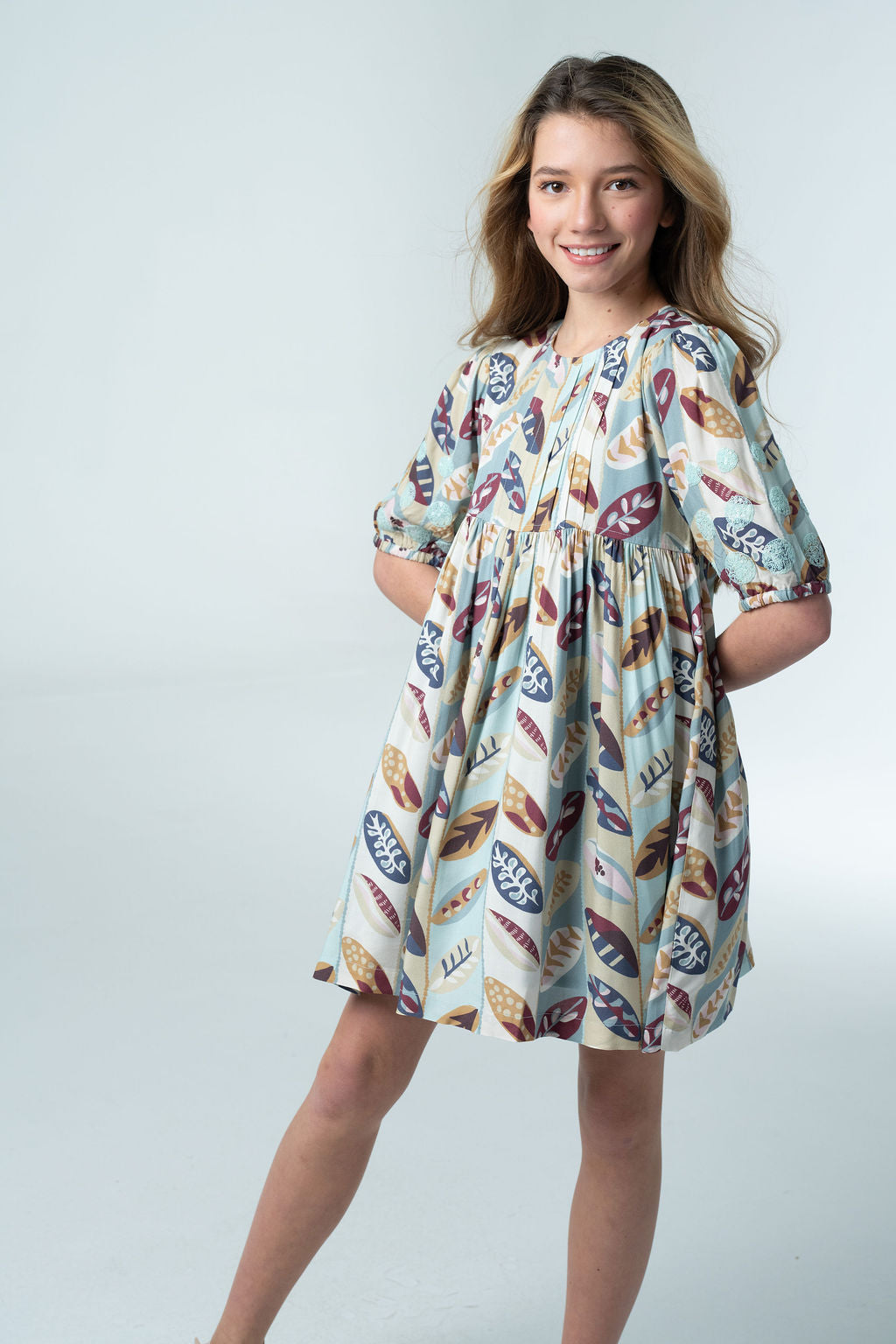 Girl wearing the Briton Court Eva Dress in the Falling Leaves print, featuring an allover leaf pattern in blue, burgundy, mustard, and sage with an A-line silhouette and puff sleeves.