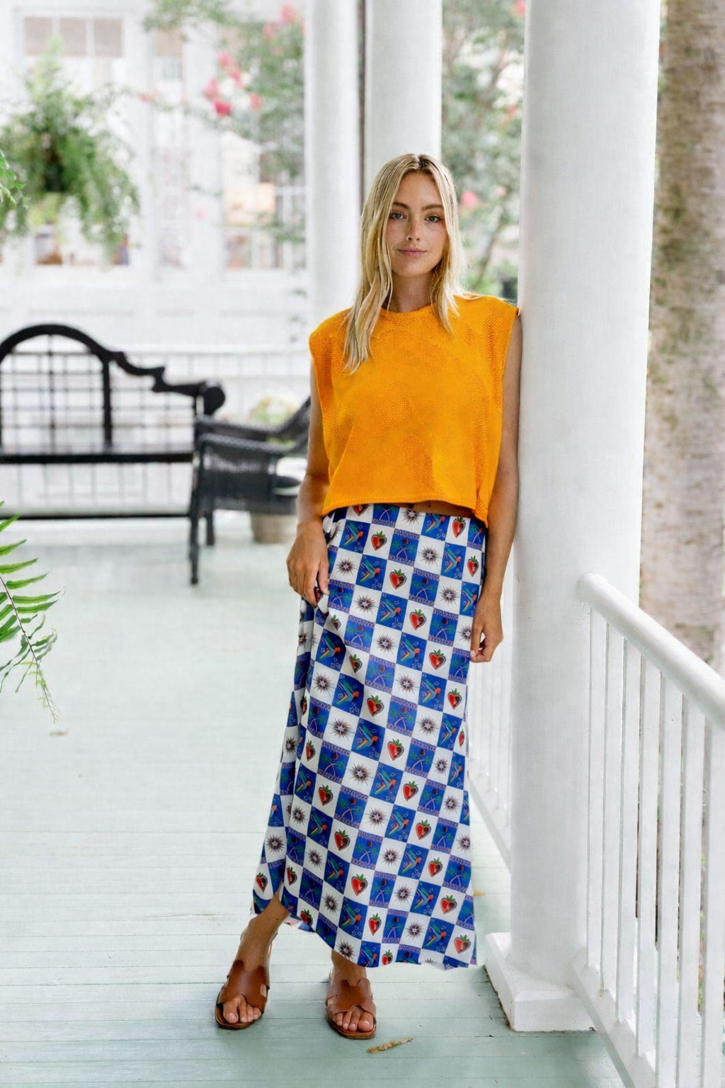 Woman wearing an orange sleeveless top and a long, patterned skirt on a porch.